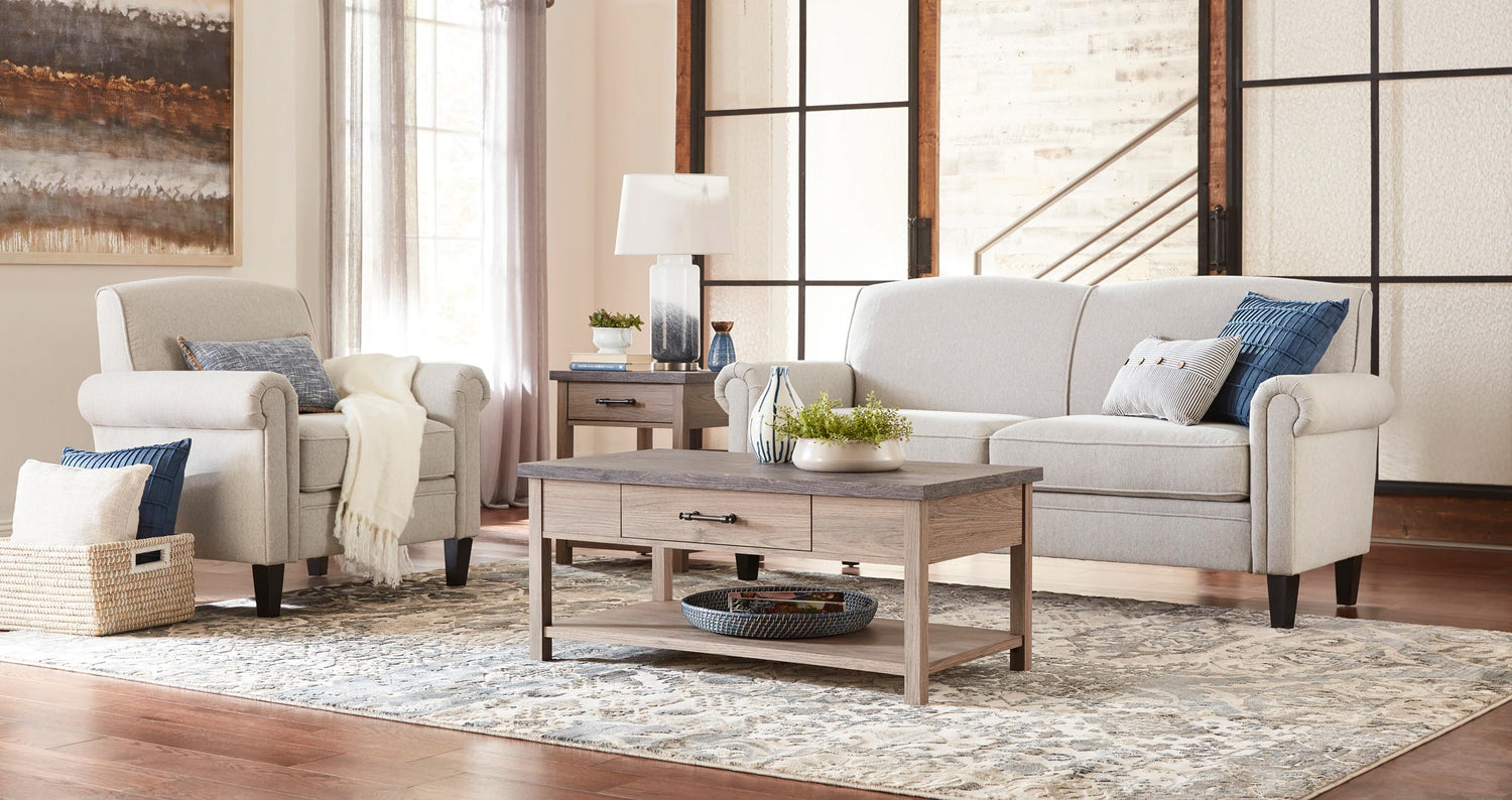A cozy living room features a beige armchair with a blue pillow, a matching beige sofa with blue cushions, and a wooden coffee table with storage. A table lamp and potted plants add decor on the side table. Light streaming through tall windows brightens the area.