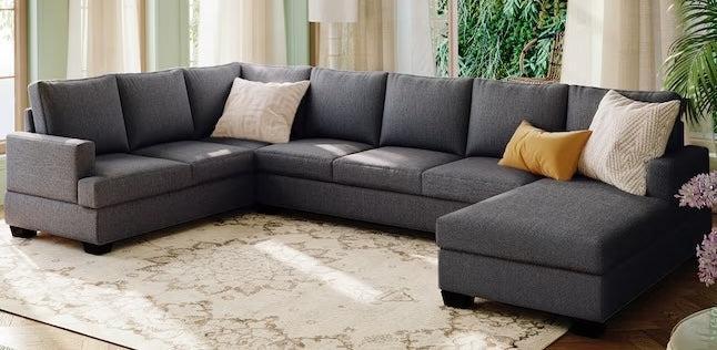 A modern, dark gray sectional sofa is arranged in a spacious, sunlit living room. The sofa is accented with a white and a yellow throw pillow. The room features large windows with curtains, a light-colored rug, and greenery visible outdoors.