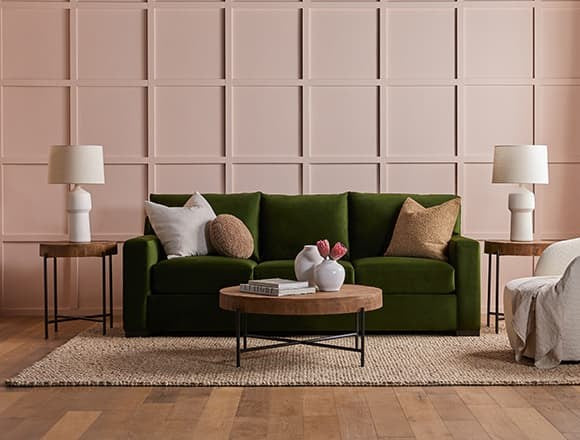 A living room with a green couch adorned with white and brown pillows, flanked by two wooden side tables with white lamps. In front, a round wooden coffee table with books and a vase of flowers sits on a beige rug. The background features a pink paneled wall.