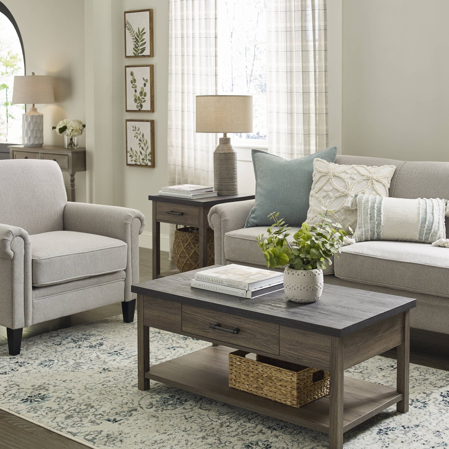 A cozy living room features a beige armchair, a light gray sofa with patterned pillows, a wooden coffee table with a drawer and shelf holding a woven basket and a plant, a side table with a lamp, and three botanical prints on the wall. Sunlight filters through sheer curtains.