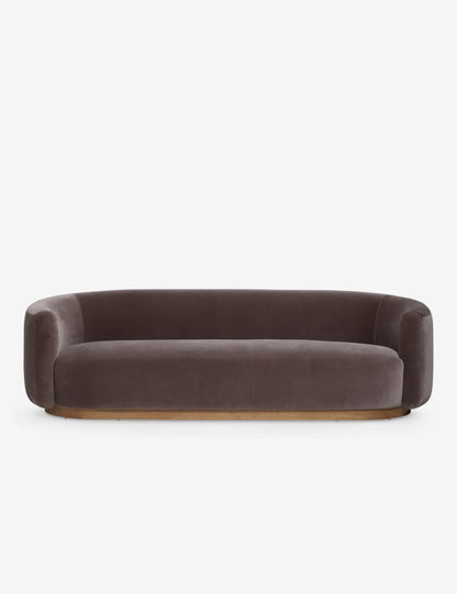 The Lowry Sofa is a stylish piece with its dark gray velvet upholstery, rounded arms, and low backrest. Set against a plain white background, it boasts a sleek, minimalist design with a wooden base that gives it an elegant and contemporary appearance.