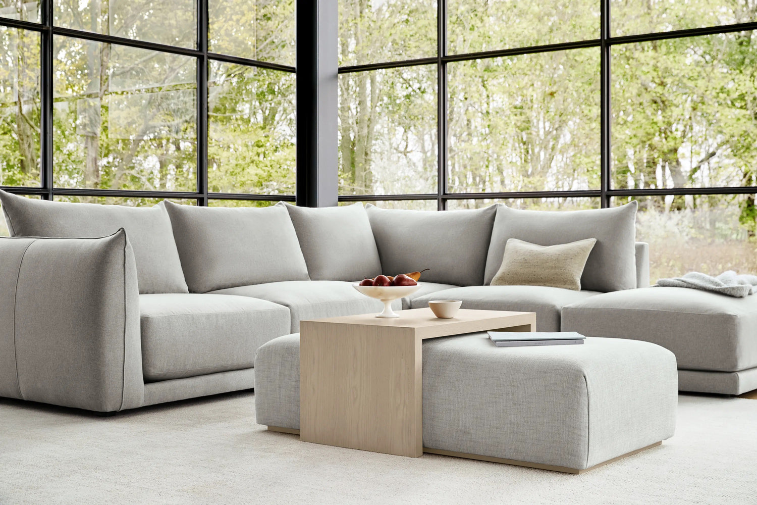 A spacious living room with large floor-to-ceiling windows showcasing a view of leafy trees. The room features a light gray sectional sofa, a matching ottoman, and a minimalist wooden coffee table. Simple d√©cor includes a small bowl of fruit and a book.