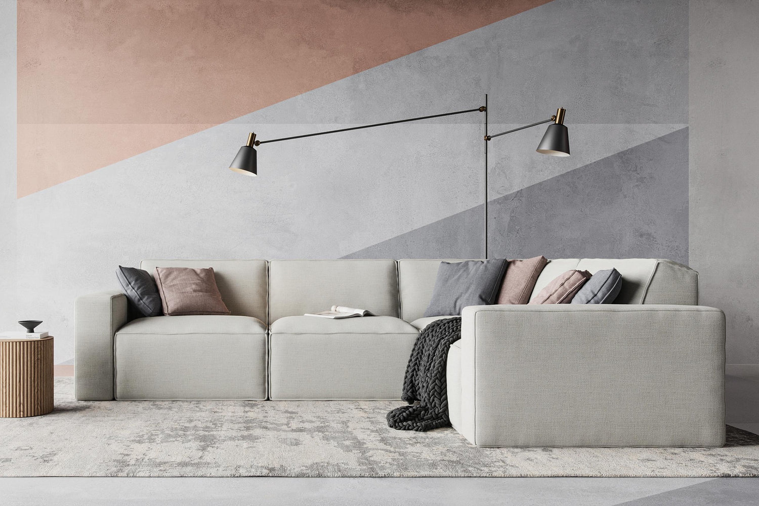 A modern living room with a light gray sectional sofa adorned with assorted pillows and a dark throw blanket. A minimalist floor lamp with two lights extends over the sofa. The wall features geometric patterns in muted tones, and a small side table stands nearby.