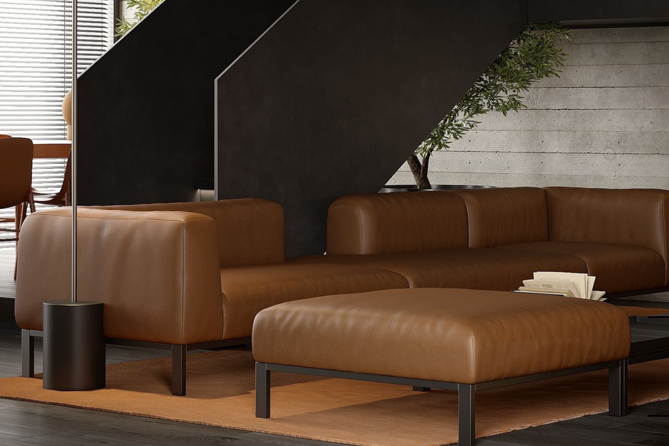A modern living room with brown leather sectional sofas and a matching ottoman on a light brown rug. The background features a concrete wall, a dark staircase, and a potted tree, with natural light streaming through blinds.