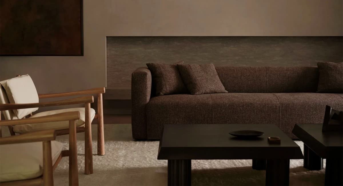 A cozy living room features a dark brown sectional sofa with two matching pillows, two wooden armchairs with white cushions, and two dark brown coffee tables. The room's neutral tones create a warm and inviting atmosphere.