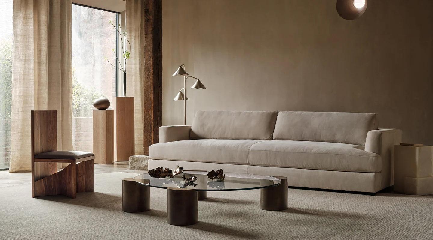 A minimalist living room featuring a beige sofa, a round glass coffee table with cylindrical legs, and a wooden chair with a modern design. Floor-to-ceiling curtains cover large windows, and a unique standing lamp with multiple light fixtures stands in the corner.