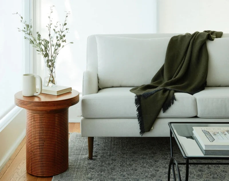 A minimalist living room showcases a white sofa adorned with a dark green throw, positioned next to a round wooden side table with a white mug, book, and a vase with foliage. A glass coffee table sits nearby on a patterned gray rug. Bright natural light fills the space.