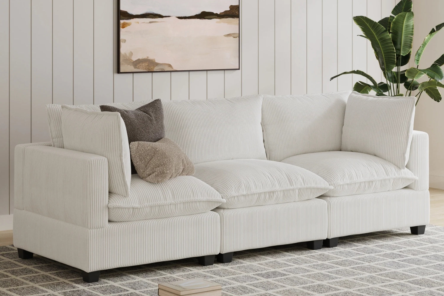 A white, L-shaped corduroy sectional sofa with multiple cushions is set against a paneled wall. A landscape painting hangs above. A green plant is on the right, and a patterned rug lies on the floor.