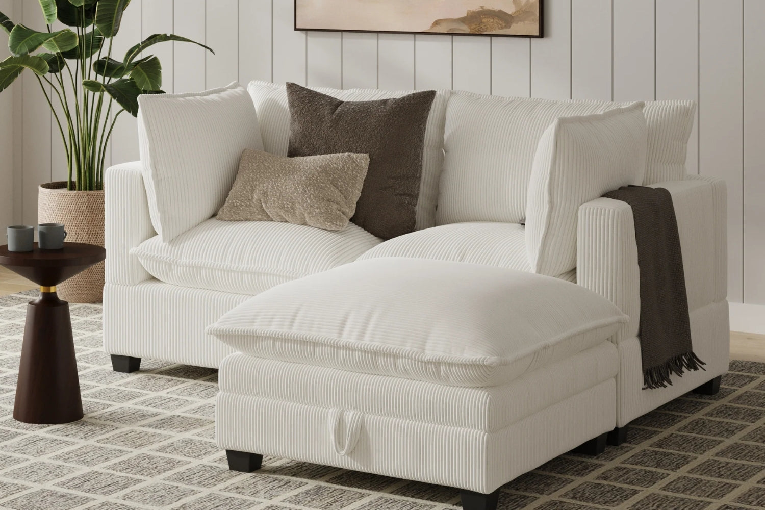 A cozy white corduroy sectional sofa with multiple cushions and a throw blanket on the right armrest. A small wooden side table with two cups is nearby. The room features light wood paneled walls, a patterned rug, and a potted plant.