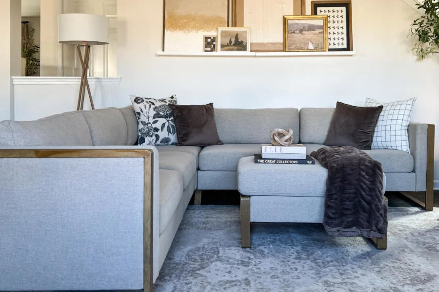 A modern living room features a light gray sectional sofa with dark and patterned throw pillows. There's a textured throw blanket on the ottoman, art pieces on a ledge, a floor lamp, and a plush area rug with subtle patterns.