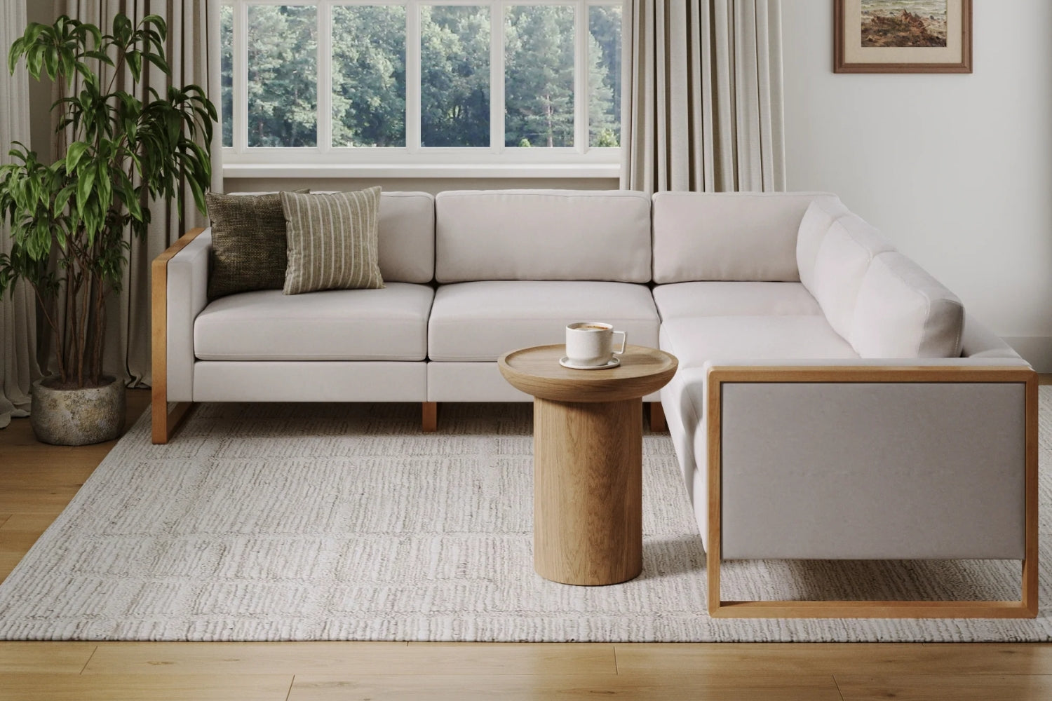 A modern living room with a light gray L-shaped sofa and a small wooden side table holding a mug. A large window reveals a view of trees. The room features a textured carpet, potted plant, and framed artwork on the wall.