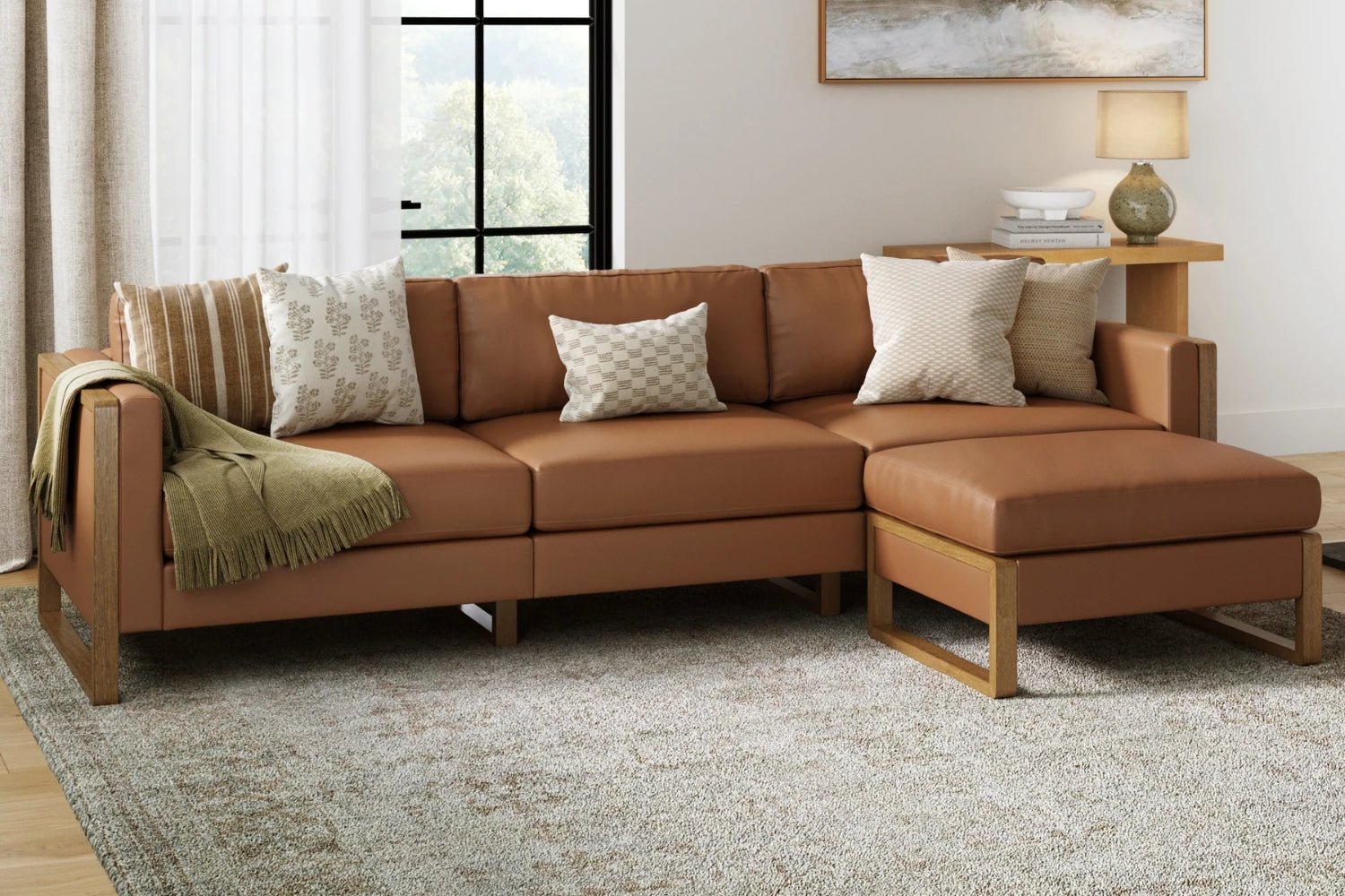 A brown sectional sofa with beige and patterned throw pillows is set in a cozy living room. A green throw blanket rests on one side. There's a wooden side table with a lamp and framed artwork. A large window and light curtains complete the scene.