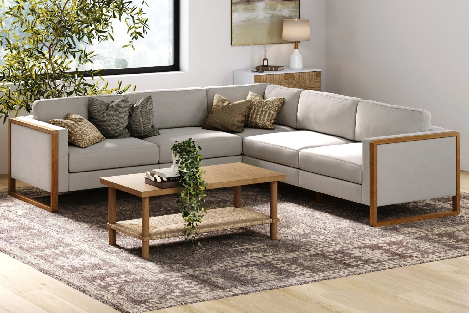 A modern living room with a beige sectional sofa adorned with patterned cushions. A wooden coffee table with a potted plant on it sits on a patterned rug. A large window and a leafy plant are in the background.
