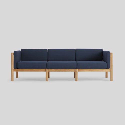 The Haven Sofa is a contemporary three-seater featuring a wooden frame and dark blue cushions. Its minimalist design emphasizes clean lines and the striking contrast between the natural wood and rich blue upholstery, set against a plain light gray background.