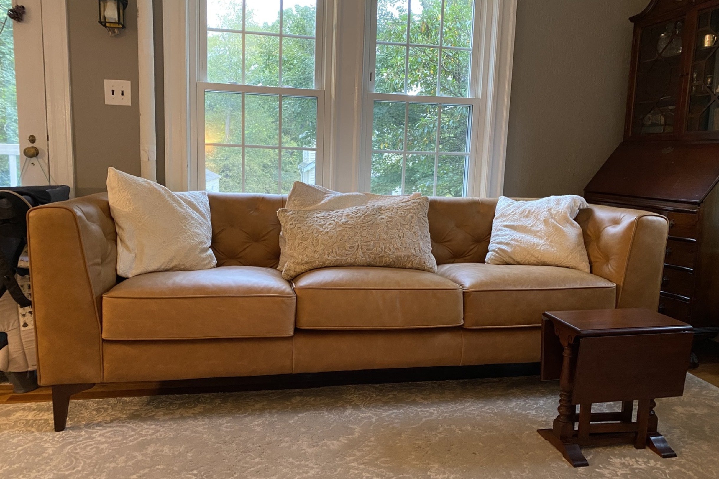 A light brown leather sofa with three large, white textured cushions is positioned in front of three large windows. A small wooden side table is placed to the right of the sofa. The room is softly lit, and greenery is visible outside the windows.