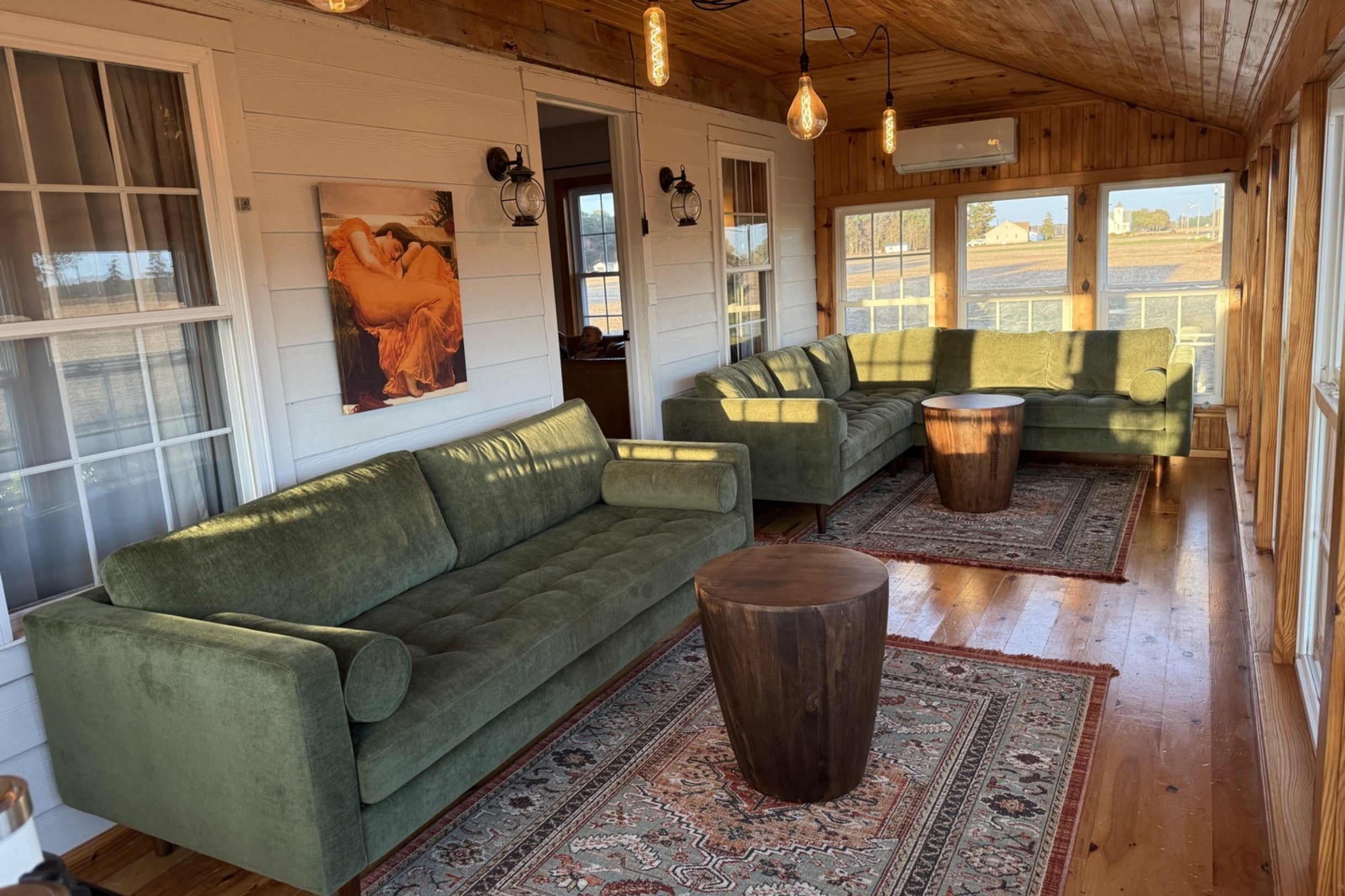 A cozy sunroom with two green sofas on patterned rugs, a wooden ceiling, and warm lighting. Two wooden stump tables are in front of the sofas. A painting of a person in orange hangs on the white wall. Large windows offer a view outside.