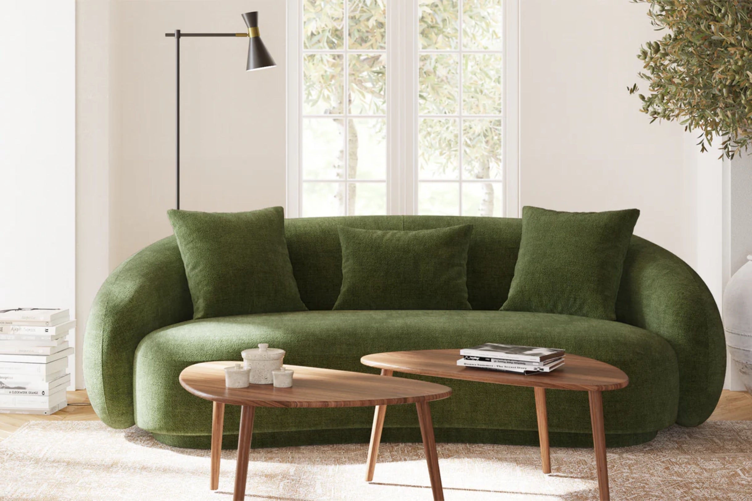 A modern living room features a green upholstered sofa with three cushions. In front are two wooden coffee tables with books and a decorative item. A tall floor lamp and a stack of books are to the side. Large windows and plants complete the scene.