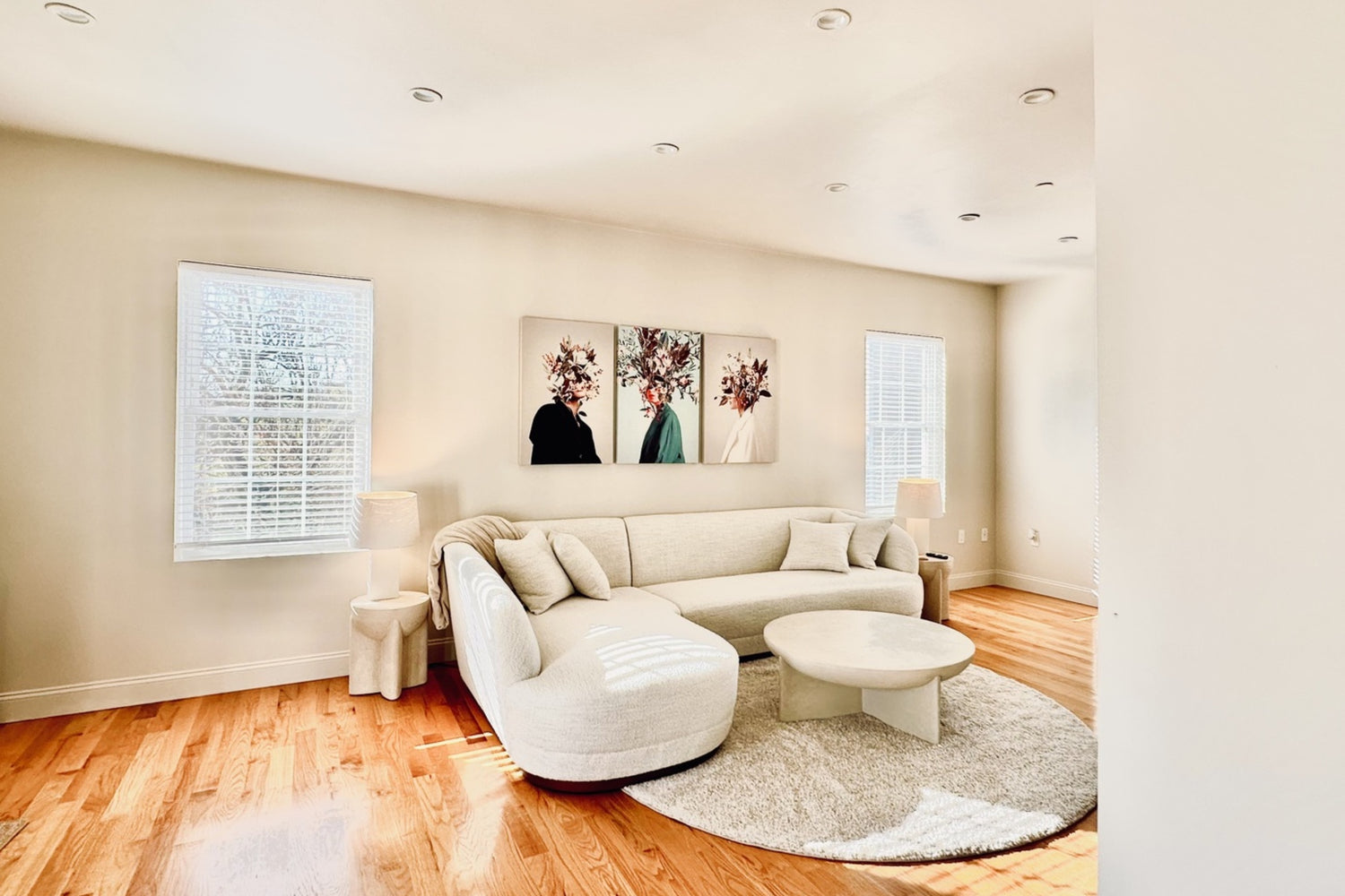 A bright living room with light wooden floors features a curved white sofa, a round white coffee table on a circular rug, and two windows with blinds. The walls display floral artwork, and there are matching lamps on side tables.