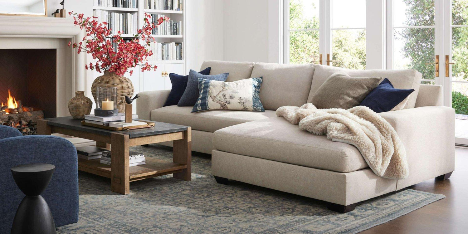 A cozy living room features a light beige sectional sofa adorned with decorative pillows and a plush blanket. A dark wooden coffee table sits in front of it, displaying a vase with red flowers and other decor items. A fireplace and bookshelf are visible in the background.