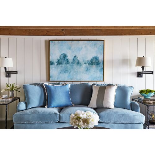 A cozy living room features the Brooke 6-Seat Sofa adorned with pillows in shades of blue and beige. An abstract blue painting hangs on a white shiplap wall above the sofa. Side tables showcase plants and lamps, while a vase with flowers rests on the coffee table.