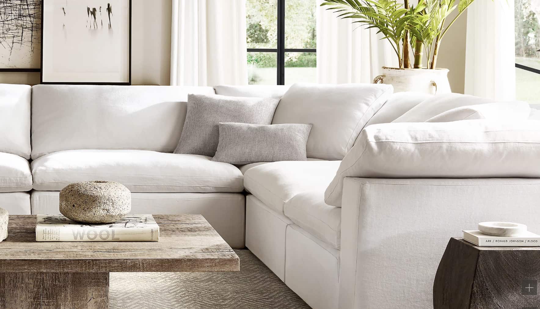 A bright and airy living room featuring a white sectional sofa with a grey accent pillow, a rustic wooden coffee table adorned with books and a stone sculpture, and a potted plant beside a large window with white curtains.