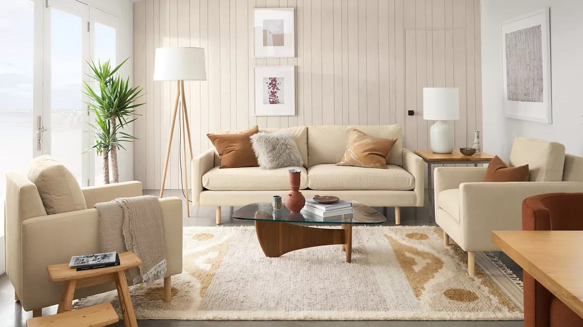 A modern living room features a beige sofa with assorted throw pillows, two matching armchairs, and a wooden coffee table with a glass top. A tall floor lamp and a large potted plant are in the corners. The room is decorated with wall art and a patterned rug.