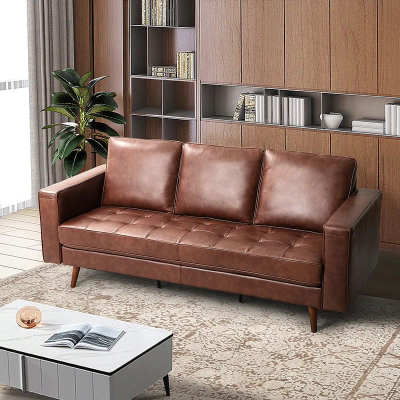 A modern living room featuring the Anatole Leather Sofa in a rich brown leather, complemented by a light gray coffee table adorned with a book and decorative piece. The space is anchored by a plush beige rug, while wooden bookshelves filled with books add warmth. A green plant near a window dressed in white curtains completes the inviting setting.