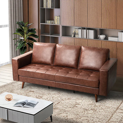 A modern living room featuring the Anatole Leather Sofa in a rich brown leather, complemented by a light gray coffee table adorned with a book and decorative piece. The space is anchored by a plush beige rug, while wooden bookshelves filled with books add warmth. A green plant near a window dressed in white curtains completes the inviting setting.
