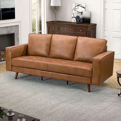 The Anatole Leather Sofa, featuring three cushions and a brown leather finish, is placed in the living room. Behind it stands a wooden sideboard adorned with decorative items and a lamp. The room includes a fireplace and art on the walls, complemented by a gray area rug and coffee table.