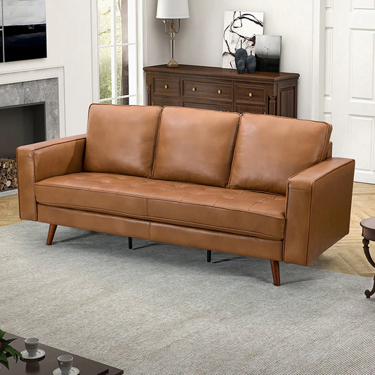 The Anatole Leather Sofa, featuring three cushions and a brown leather finish, is placed in the living room. Behind it stands a wooden sideboard adorned with decorative items and a lamp. The room includes a fireplace and art on the walls, complemented by a gray area rug and coffee table.