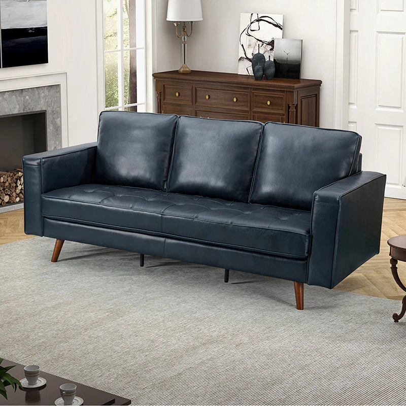 A modern living room features the Anatole Leather Sofa in dark blue, resting on a light gray rug. Behind the sofa, there is a wooden sideboard adorned with decorations and a white table lamp. To the left, a fireplace with stacked logs adds warmth to the space, while a small coffee table sits in the foreground.
