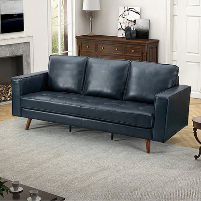 A modern living room features the Anatole Leather Sofa in dark blue, resting on a light gray rug. Behind the sofa, there is a wooden sideboard adorned with decorations and a white table lamp. To the left, a fireplace with stacked logs adds warmth to the space, while a small coffee table sits in the foreground.