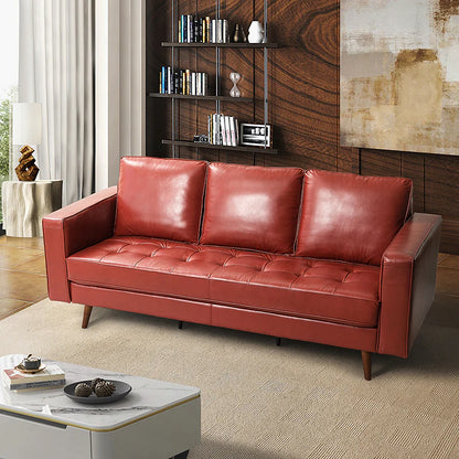 An Anatole Leather Sofa in red, featuring three cushions, is situated in a modern living room. The background includes a bookshelf and a piece of textured wall art. A small coffee table with a decorative centerpiece is placed in the foreground, while light curtains frame the scene.