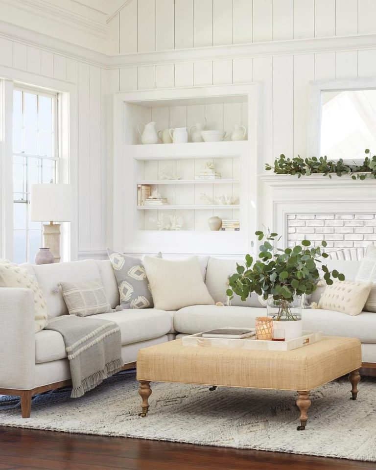A bright and airy living room features a light gray sectional sofa with patterned cushions, a textured beige ottoman, and a lush green plant centerpiece. White shiplap walls, built-in shelves with decorative items, and a brick fireplace complete the cozy space.
