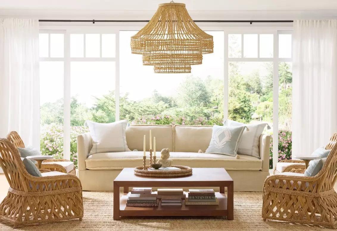 A bright, airy living room featuring a beige sofa with white and blue cushions, surrounded by four wicker chairs. A wicker chandelier hangs from the ceiling, and a wooden coffee table sits in the center, adorned with candles and decor. Large windows offer a view of greenery outside.