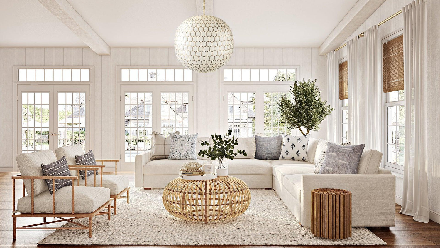 A bright living room with large windows, white walls, and light wood floors. It features cream-colored sofas, a round wicker coffee table, a large plant, and a textured rug. A modern pendant light hangs from the ceiling. The windows are adorned with sheer curtains and bamboo blinds.
