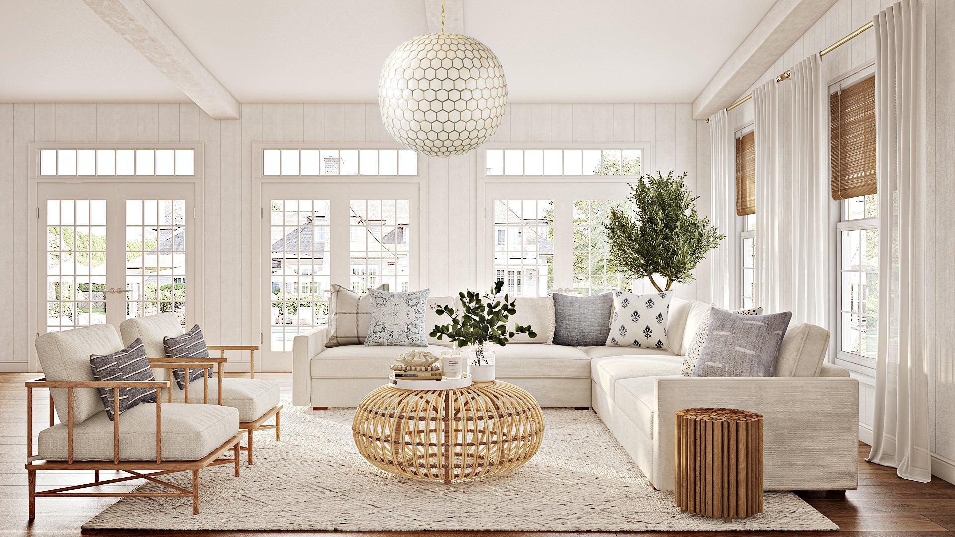 A bright living room with large windows, white walls, and light wood floors. It features cream-colored sofas, a round wicker coffee table, a large plant, and a textured rug. A modern pendant light hangs from the ceiling. The windows are adorned with sheer curtains and bamboo blinds.