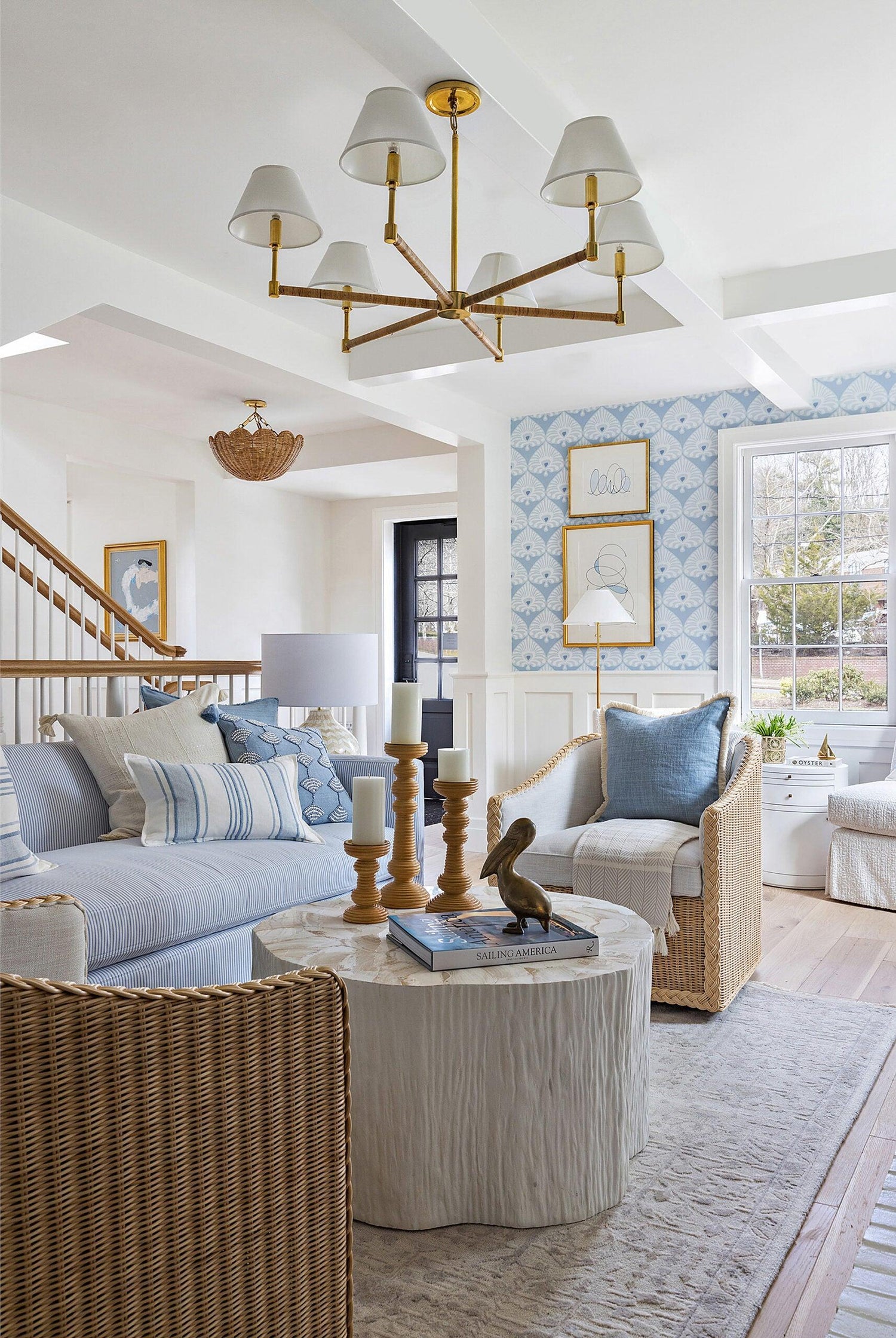 A stylish living room with a light blue and white color scheme. It features a light blue sofa with striped and patterned pillows, wicker chairs, and a coffee table with candles and books. A chandelier hangs from the ceiling. The walls are decorated with framed art.