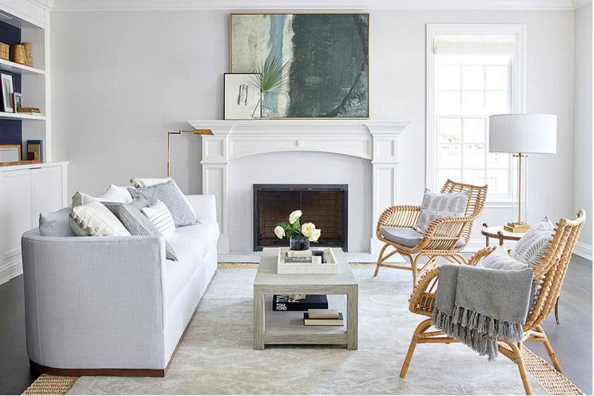 A modern living room with a light gray sofa, two wicker chairs with gray cushions, and a white fireplace. A large abstract painting hangs above the fireplace. A coffee table is placed in the center with decorative items, and a floor lamp stands near the window.