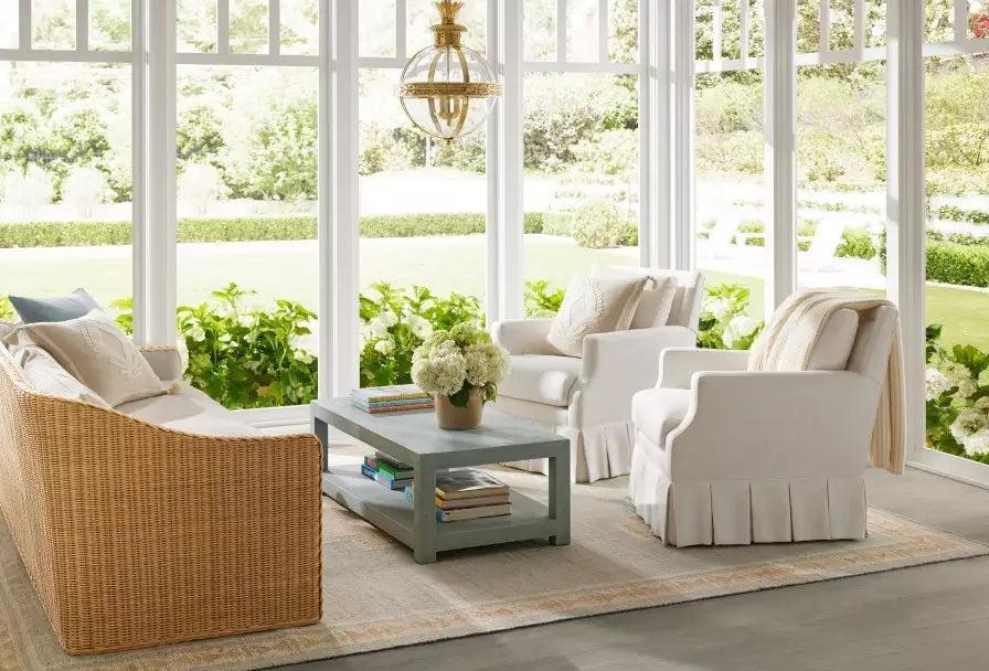 A bright sunroom with large windows overlooking a lush garden. The room features a wicker sofa, two white armchairs, a wooden coffee table with books and a vase of flowers, and a neutral-toned rug. The space is adorned with greenery and a hanging light fixture.