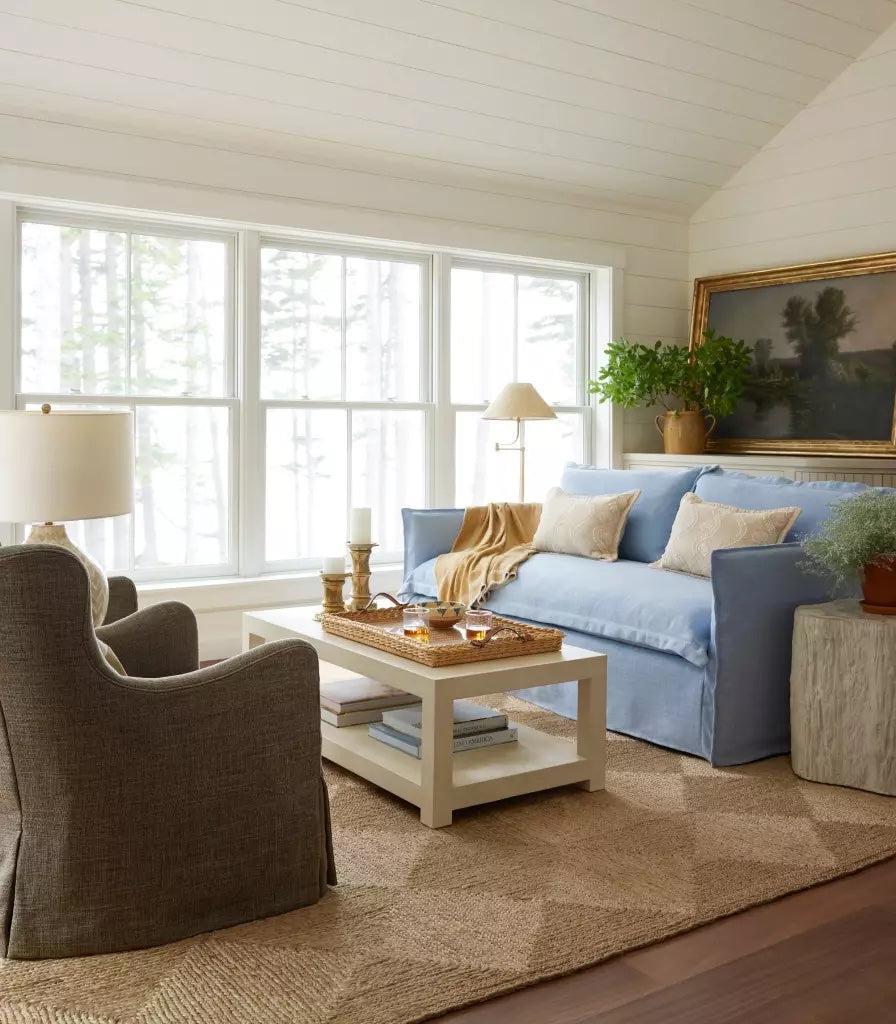 A cozy living room with a large window receiving natural light. It features a blue sofa with white pillows, a beige armchair, a white coffee table with decor items, a potted plant, a floor lamp, and wall art. The walls and ceiling are white, with a textured beige rug on the floor.