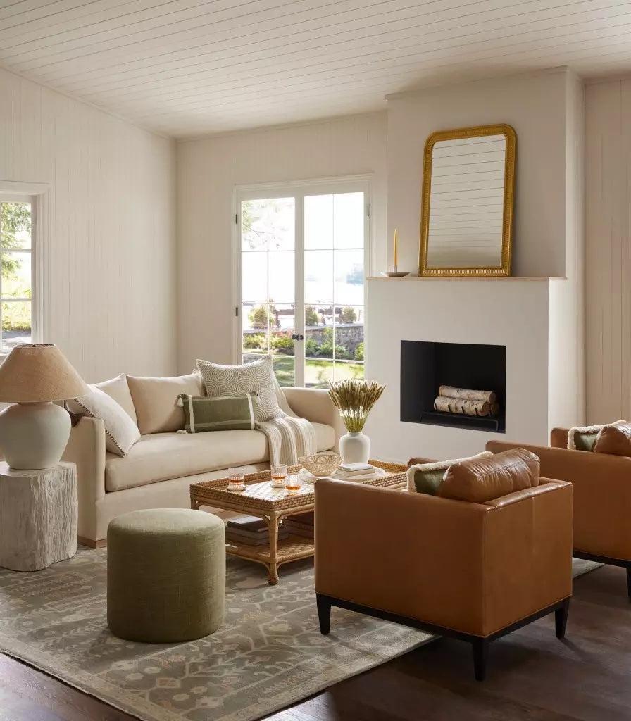 A cozy living room with a beige sofa, two tan leather chairs, and a green ottoman arranged around a wicker coffee table on a patterned rug. A fireplace with a gold-framed mirror above and large windows letting in natural light complete the space.