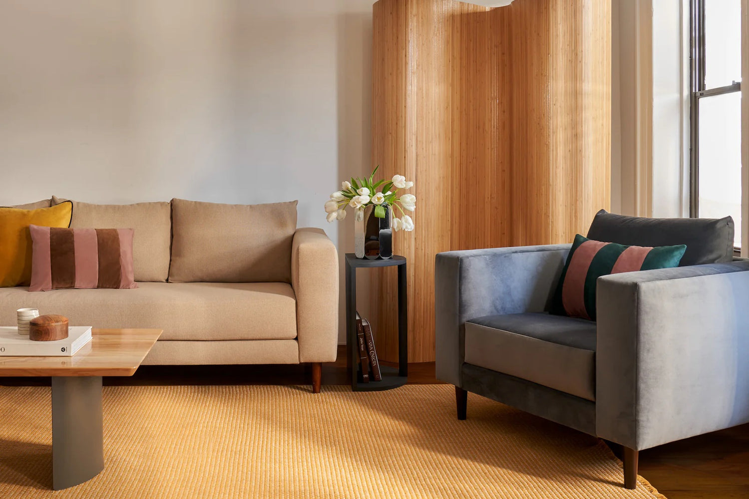 A modern living room with a beige sofa and a blue armchair, both featuring colorful throw pillows. A wooden coffee table stands on a yellow rug, and a small side table with a vase of white flowers is placed beside the sofa. A wooden room divider is in the background.