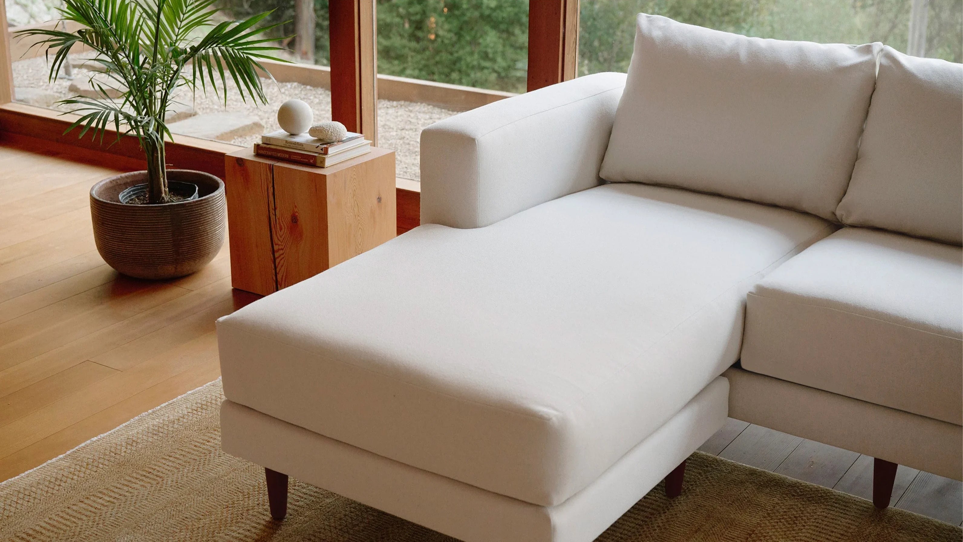 A modern white sectional sofa with a chaise lounge is positioned near a large window. A wooden side table holding decorative items and a potted plant sits beside the sofa. The room has a warm feel with natural light, wooden flooring, and a beige rug.