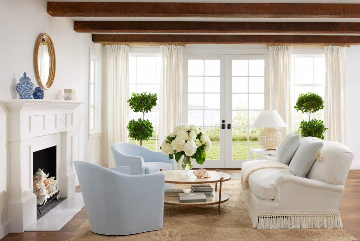 Bright living room with white walls, wooden ceiling beams, and large windows with a view of the outdoors. The room features a white sofa, two light blue armchairs, a round wooden coffee table, a fireplace adorned with decorative items, and lush greenery.
