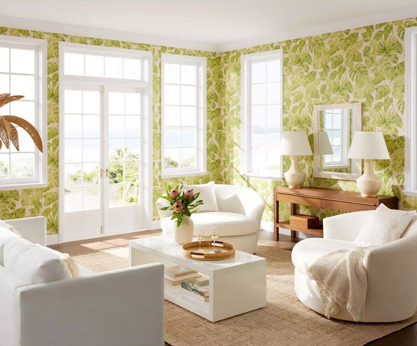 A bright living room with tropical green leaf-patterned wallpaper, large windows, and glass doors allowing natural light. The room features white, modern furniture including sofas, armchairs, a coffee table, and a wooden console table with two lamps. A vase of flowers is on the coffee table.