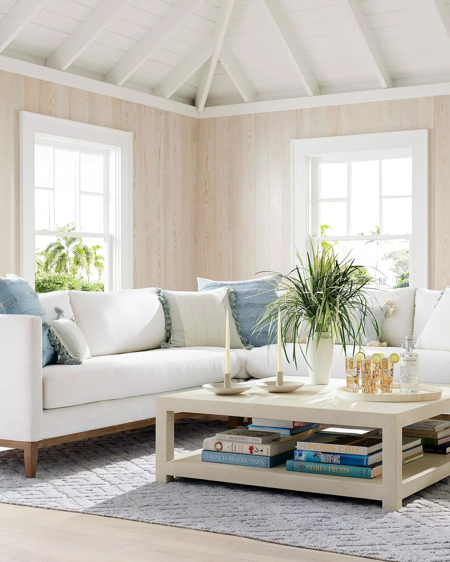 A bright, airy living room with white walls and high, angled ceilings. A white sectional sofa with blue and white pillows faces a wooden coffee table, which is adorned with a plant, candles, and books. Sunlight streams through two large windows, highlighting a cozy ambiance.