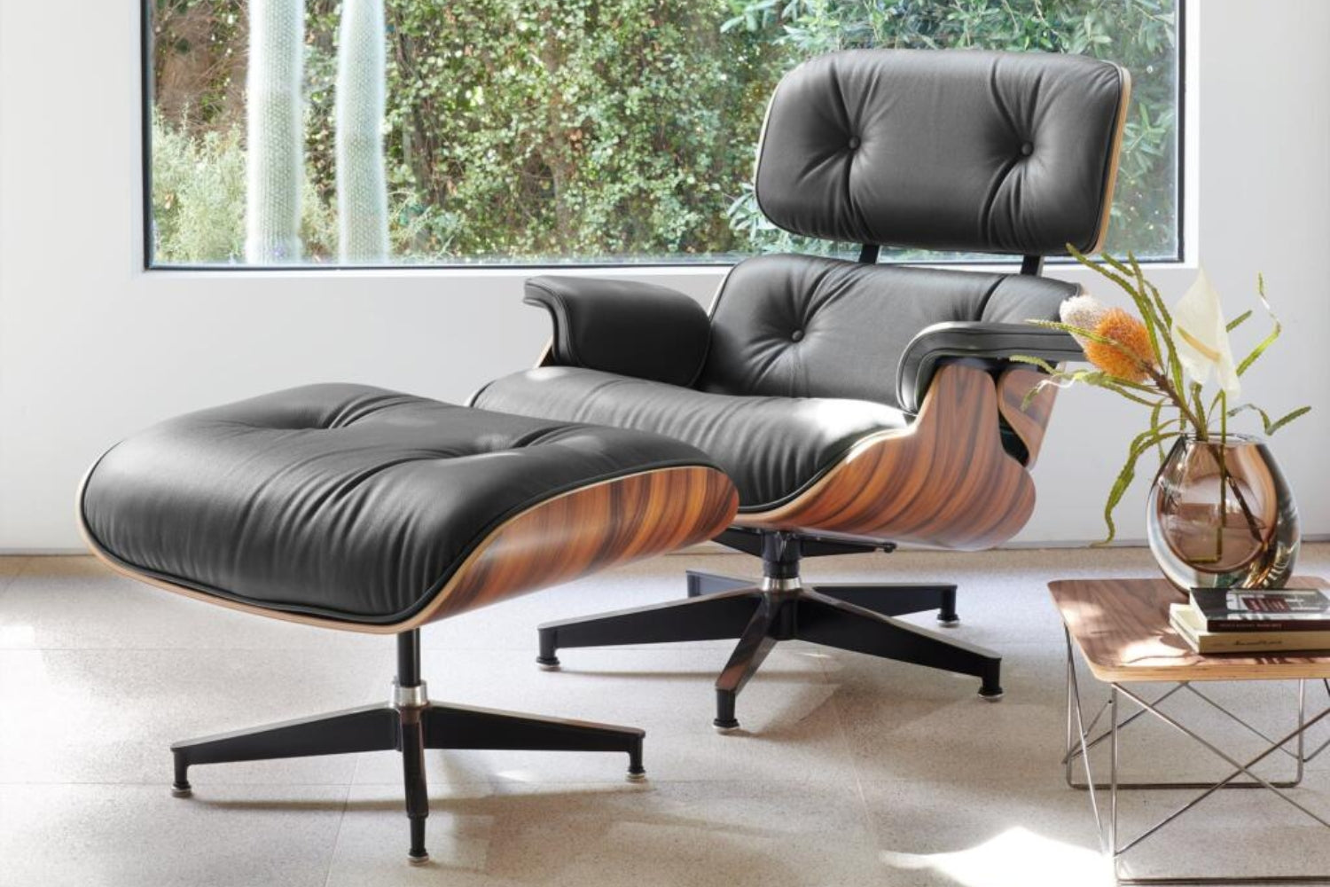 A modern black leather lounge chair with a matching ottoman is placed in a well-lit room. The chair has wooden accents and metal legs. Nearby, there's a small table with decorative plants and a large window showcasing greenery outside.