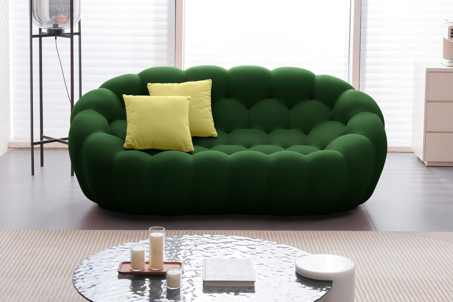 A modern living room with a dark green, tufted sofa and two yellow cushions. A round coffee table with candles and a small bowl is in the foreground, and large windows are in the background.