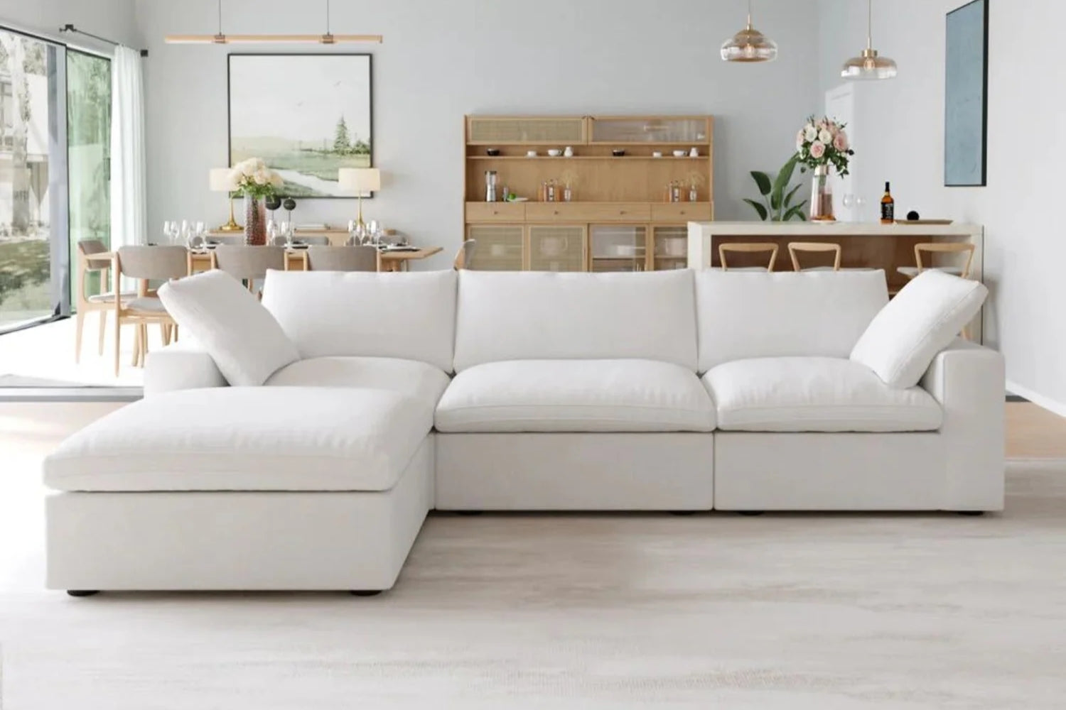 A modern living room with a large, white sectional sofa. Behind the sofa, a dining table is set with chairs and floral arrangements. Hanging lights illuminate the area. Large windows on the left let in natural light. Minimalist decor throughout.
